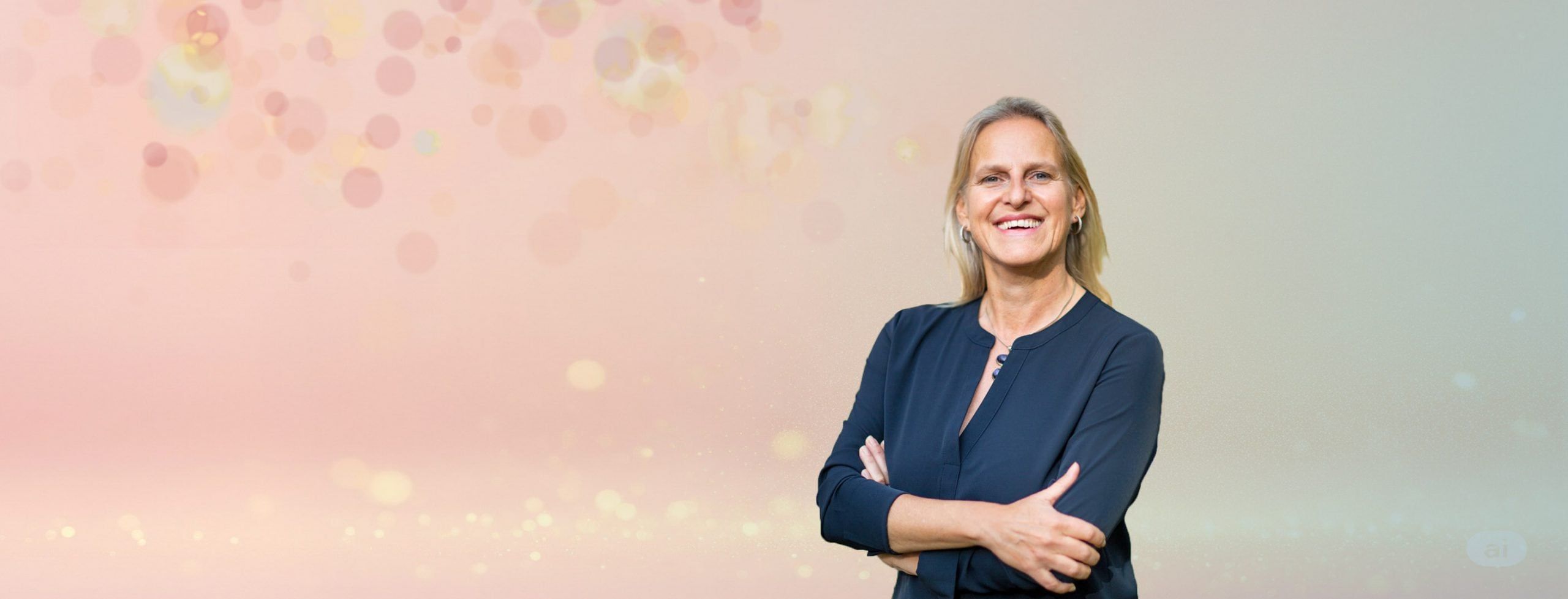 Nicole Rupp lächelt mit verschränkten Armen vor einem hellen, rosa schimmernden Hintergrund